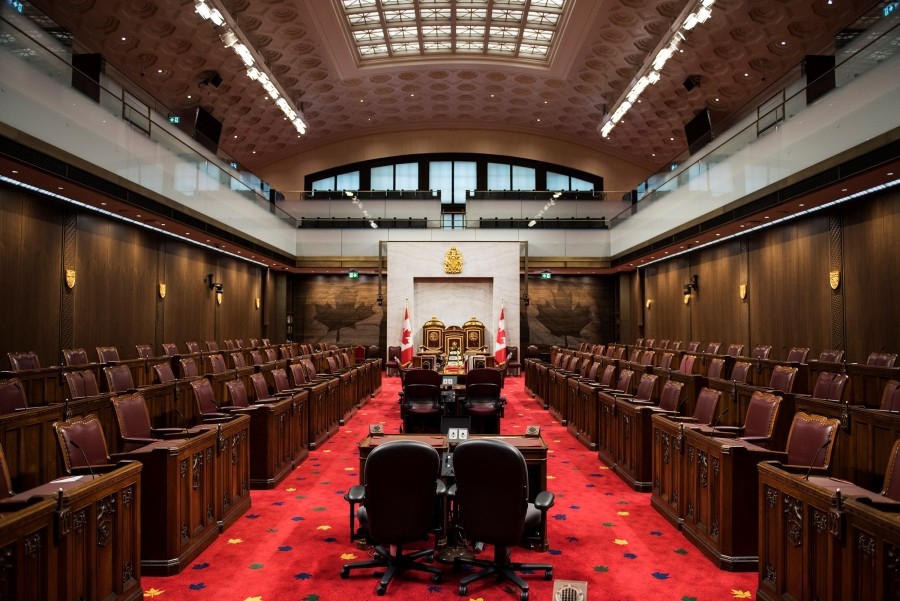 Large room with dark wood paneling and seats along both sides and a central aisle with desks and red carpet.